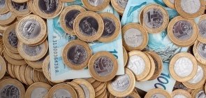 Foto mostra duas notas de cem reais e diversas modas de um real estão espalhadas sobre uma mesa.