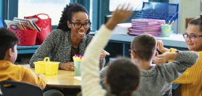 Foto de uma professora em uma mesa com grupo de crianças. Uma delas está com a mão levantada