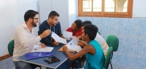 Mesa com três crianças e dois adultos, eles conversam com papeis na mão