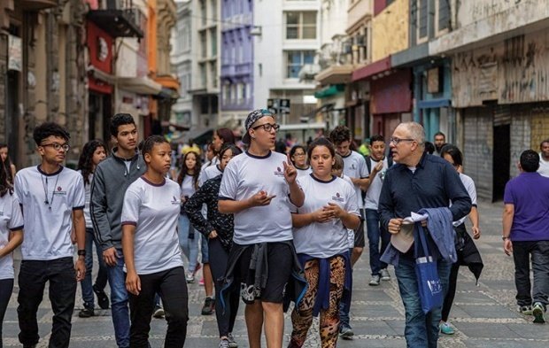 No centro de São Paulo, um grupo de alunos caminha com o professor. Destaca-se um dos alunos conversando com o professor