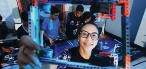 Aluna em aula de robótica segura peça construida na aula de robótica. Ela usa óculos, sorri e está com o cabelo preso