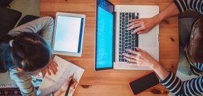 Foto de cima de uma mesa em que há uma criança sentada de um lado escrevendo em uma folha de papel e uma mulher adulta do outro, mexendo no computador