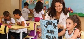 Alunos se dividem pela biblioteca, no fundo alguns escolhem livros, outros leem. No destaque a professora conversa com uma aluna que segura um livro escrito 
