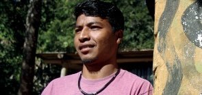 Homem indígena está usando camiseta rosa, ele olha para o lado e está com os braços cruzados