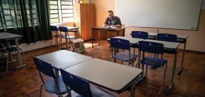 Professor senta sozinho em uma sala de aula, ele aparece no fundo lendo sentado, usa camiseta preta e óculos