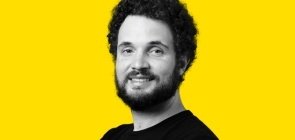 Fundo amarelo, retrato de um homem em branco e preto por volta dos 30 anos, com barba e cabelo. Ele sorri para a foto