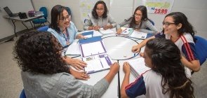 Cinco alunas prestam atenção em uma professora em uma mesa redonda. A professora segura um fichário. As alunas vestem uniforme, três delas também usam casaco cinza