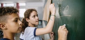 Uma menina e um menino estão na lousa resolvendo contas. Os dois vestem uniforme