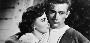 Fotografia em branco e preto. São dois jovens, o homem abraça a mulher e olha para o lado, ela olha para ele