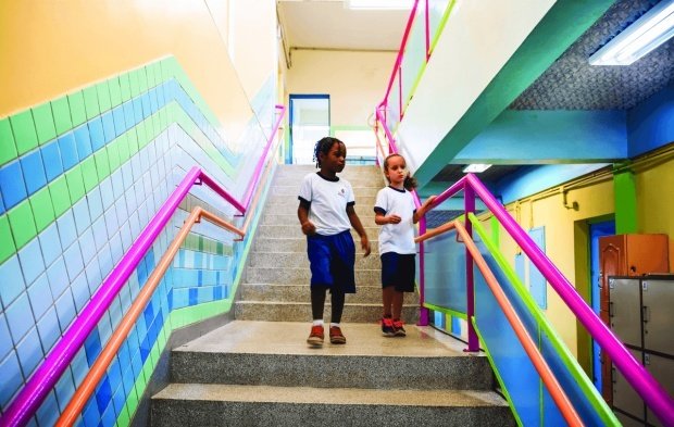 Duas meninas vestindo o uniforme descem a escada. Com as paredes e o corrimão coloridos, o teto tem forro acustico