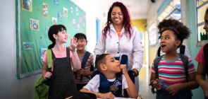 Uma professor empurra a cadeira de rodas de um aluno, que está rodeado de outros alunos. Todos sorriem. 