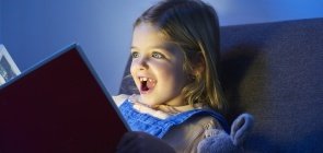 Menina loira está sentada com um livro com aberto, o rosto dela está iluminado