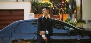 Homem veste camisa preta, ele sorri, está encostado em carro preto