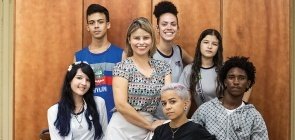 Grupo de jovens, não usam uniforme. Mulher mais velha, professora, se apoia em cadeira onde jovem senta