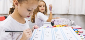 Menina sentada olhando para uma cartilha com as vogais necessárias para escrever diferentes palavras e com as consoantes faltando