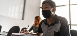 Um aluno está sentado em sala de aula e anota coisas em um caderno. Ele usa máscara. 