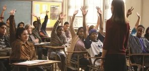 Sala de aula, alunos sentados levantam a mão, professora está de costas para a foto e olha para eles