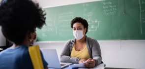 Uma professora está sentada em sua mesa dentro da sala de aula e uma aluna olha em sua direção. Ambas usam máscara.