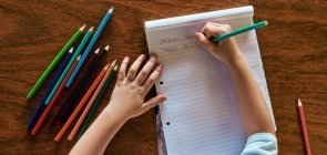 Criança com caderno aberto e dois nomes escritos sobre mesa com lápis espalhados