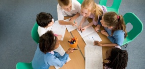 Seis crianças interagem em torno de uma mesa redonda dentro de uma sala de aula