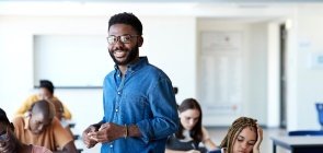 Um professor de cerca de 40 anos de pé entre carteiras da sala de aula com alunos adolescentes sentados escrevendo