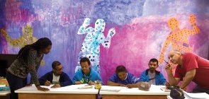 Quatro alunos sentados um ao lado do outros enquanto dois professores conversam com eles 