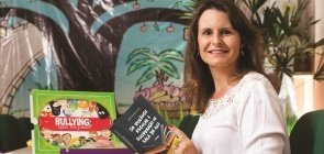 Mulher branca de cabelo médio escuro, veste uma camisa branca, está sentada e segura livro. Na mesa há livros e uma caixa verde escrita bullying
