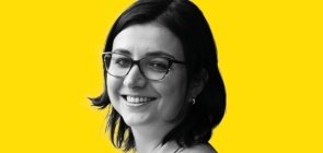 Mulher de óculos, está de lado e sorri para a foto. Ela tem cabelo na altura do ombro