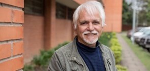 Homem branco por volta dos 60 anos, de cabelo branco. Ele veste um sueter azul marinho e uma jaqueta cinza. Ele sorri