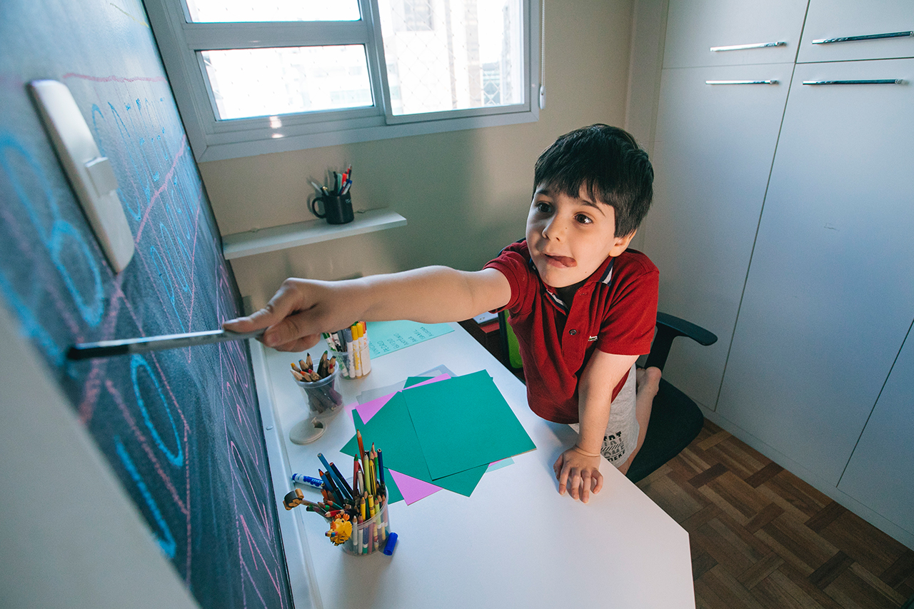 Fotografia de aluno apontando para letras que est&atilde;o marcadas na lousa a frente da mesa.