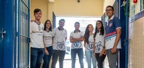 Um grupo de 7 alunos de cerca de 15 anos felizes lado a lado no portão de uma escola