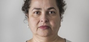 Mulher de aproxidamente 30 anos, cabelo cacheado e recolhido, olha fixamente para a câmera e olhar sério. 