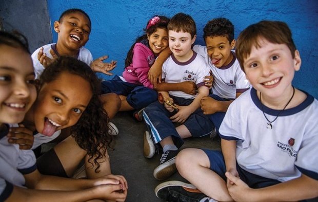 Grupo de sete alunos sentados no chão, usam uniforme da prefeitura de são paulo, sorriem para a foto