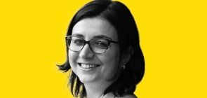 Mulher de óculos, está de lado e sorri para a foto. Ela tem cabelo na altura do ombro