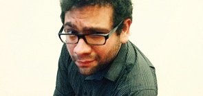 Homem de camisa listrada se aproxima da foto, ele usa oculos preto, tem braços cruzados e está em pé. Ele tem uma expressão de questionamento