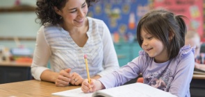 Uma professora está sentada ao lado de uma aluna que escreve em seu caderno