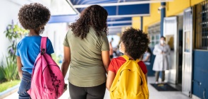 De costas, andando pelo corredor de uma escola, uma mulher caminha de braços dados com duas crianças. Elas carregam uma mochila nas costas.