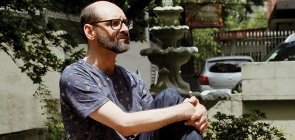 Homem sentado de lado, ele tem uma perna em pé abraçada. Ele olha para o horizonte, é careca, usa óculos, veste uma camiseta azul estampada e um moletom