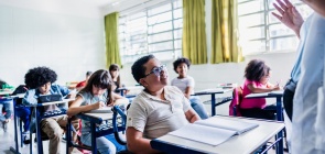 Sala de aula de alunos dos Anos Iniciais. Professora na frente dando aula. Em destaque, na primeira carteira, um aluno com deficiência