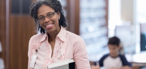 Professora, em pé, segura livros e sorri