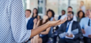 Uma mulher está a frente, em pé, em uma sala com plateia.
