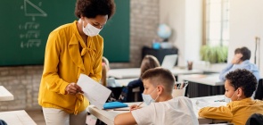 Uma professora mostra uma folha para um aluno dentro de uma sala de aula. Todos usam máscara