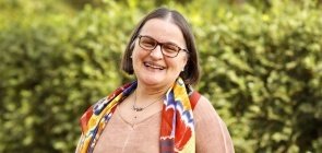 Retrato de uma senhora de aproximadamente 60 anos de idade. Ela sorri para a câmera. Tem os cabelos lisos e curtos, bastante escuros. Tem um óculos vermelho escuro, veste um lenço colorido e uma blusa da cor da pele clara. Ao fundo, folhagens.