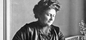 Foto de Maria Montessori trabalhando em sua mesa. Ela olha para os livros