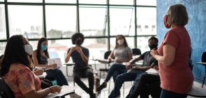 Uma professora fala em pé enquanto outros escutam atentamente em uma sala de aula