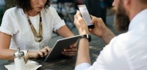 Sem mostrar o rosto, aparecem um homem e uma mulher. Ela de camiseta branca e colar marrom mexe em um tablet e ele de camisa social branca mexe no celular. Na mesa onde estão sentados está um chá gelado e um açucareiro