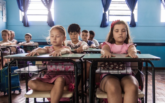 Alunos meditam em sala de aula. Eles estão com os olhos fechados e os braços esticados sobre a carteira.