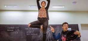 Em cima da mesa, uma menina em pé com as mãos unidas em cima da cabeça, uma perna dobrada. No fundo uma lousa com tipos de desenho e uma professora explicando 