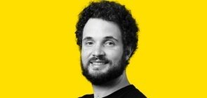 Fundo amarelo, retrato de um homem em branco e preto por volta dos 30 anos, com barba e cabelo. Ele sorri para a foto