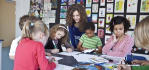 Uma professora acompanha um grupo de crianças que desenham sobre uma mesa dentro de uma sala de aula
